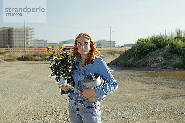 Selbstbewusste Frau hält Topfpflanze und Gießkanne auf der Baustelle