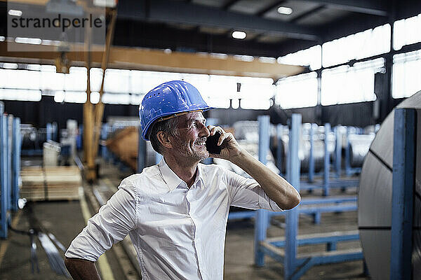 Lächelnder männlicher Ingenieur mit Helm  der im Stahlwerk mit dem Handy spricht