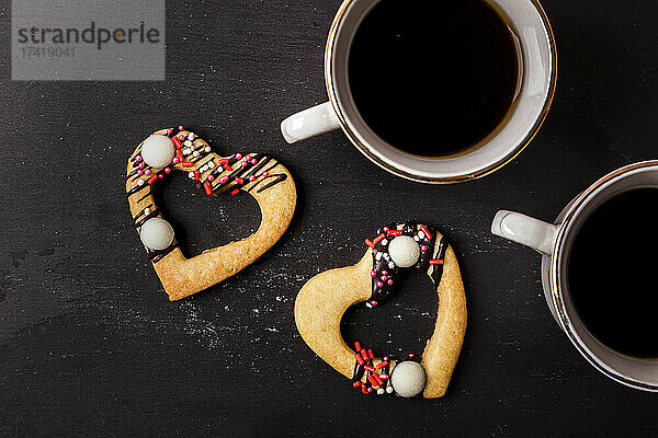 Studio shot of heart shaped cookies and two cups of coffee standing against black background