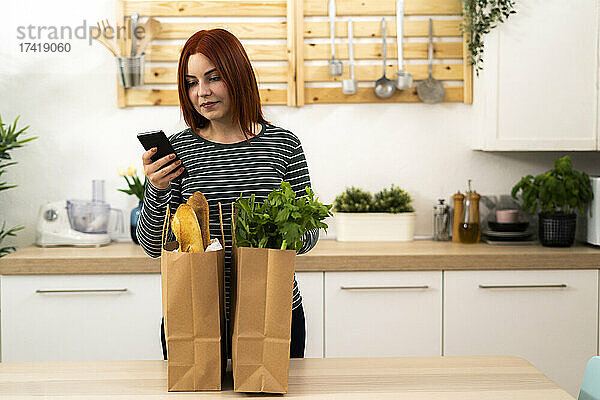 Frau benutzt Mobiltelefon  während sie mit Einkaufstüten am Tisch steht