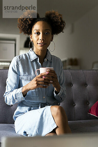 Junge Afro-Frau mit Kaffeetasse sitzt zu Hause auf dem Sofa