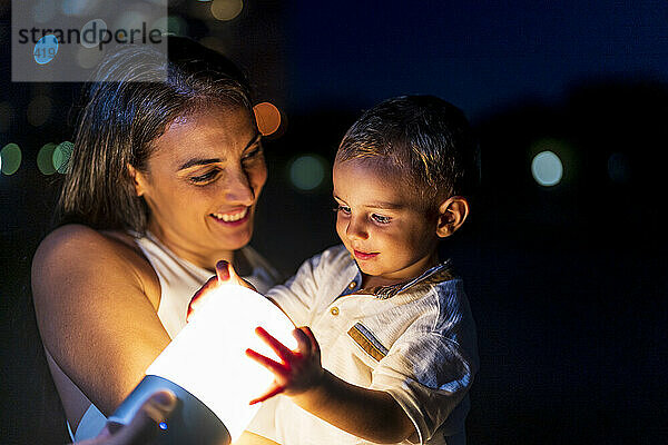 Lächelnde Frau blickt nachts auf ihren Sohn  der Beleuchtungsgeräte in der Hand hält