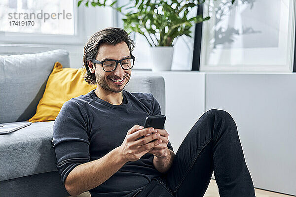Lächelnder Mann benutzt Mobiltelefon im Wohnzimmer zu Hause