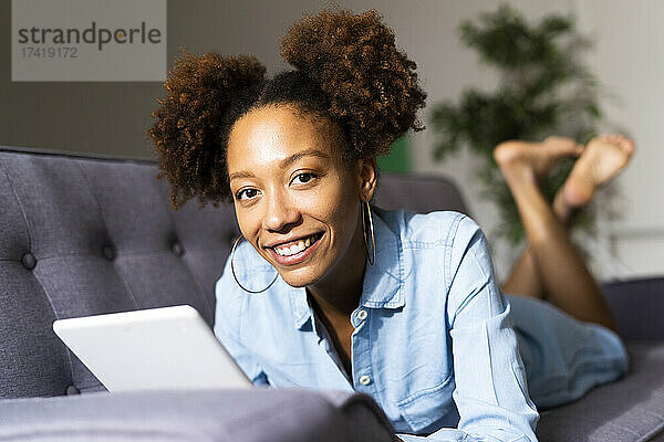 Lächelnde Afro-Frau mit digitalem Tablet liegt auf dem Sofa im Wohnzimmer