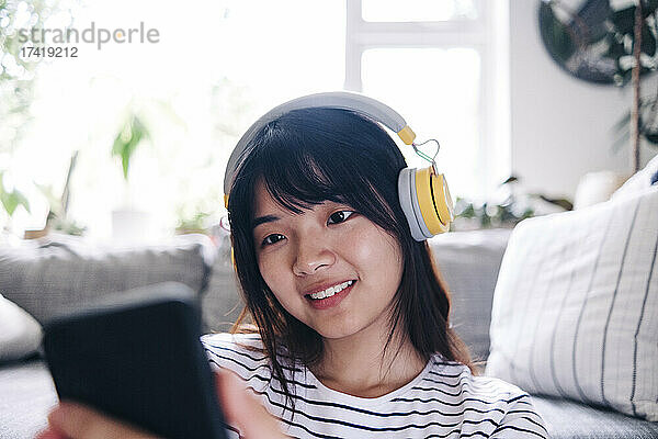 Lächelnde Frau mit kabellosen Kopfhörern  die zu Hause ihr Smartphone nutzt