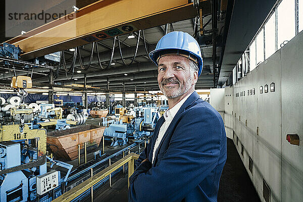 Lächelnder männlicher Ingenieur mit verschränkten Armen steht im Stahlwerk