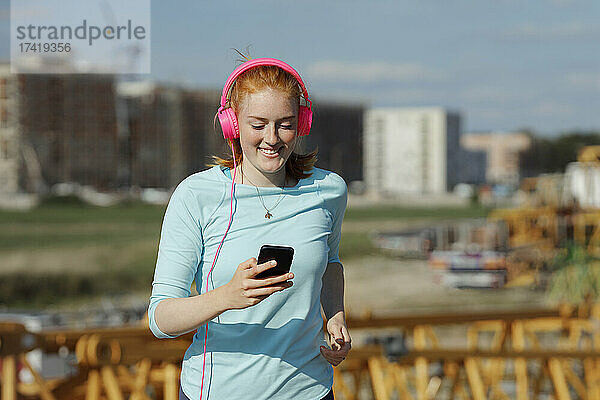 Frau mit Kopfhörern benutzt Mobiltelefon beim Joggen an einem sonnigen Tag