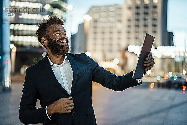 Männlicher Berufstätiger lacht  während er in der Stadt ein Selfie mit einem digitalen Tablet macht