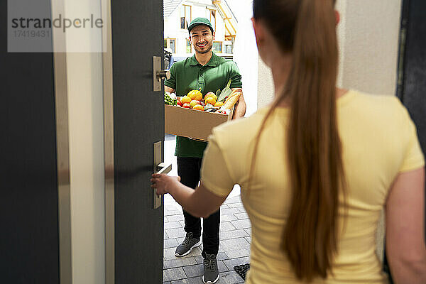 Lächelnder Lieferbote hält Gemüsekiste in der Hand  während er am Eingang steht