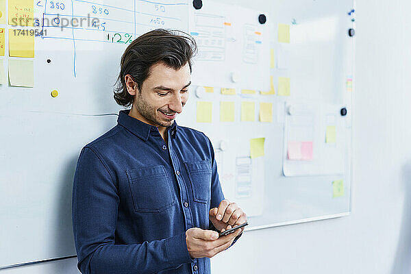 Lächelnder Geschäftsmann mit Smartphone vor Whiteboard im Büro