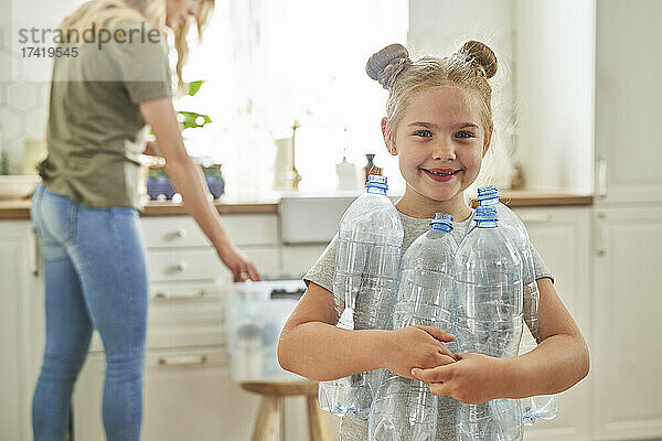 Lächelndes Mädchen hält Plastikflaschen in der Hand  während die Mutter in der Küche steht
