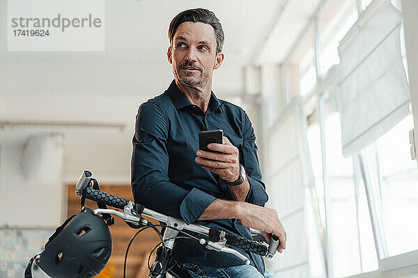 Reifer Mann mit Smartphone und Fahrrad im Büro