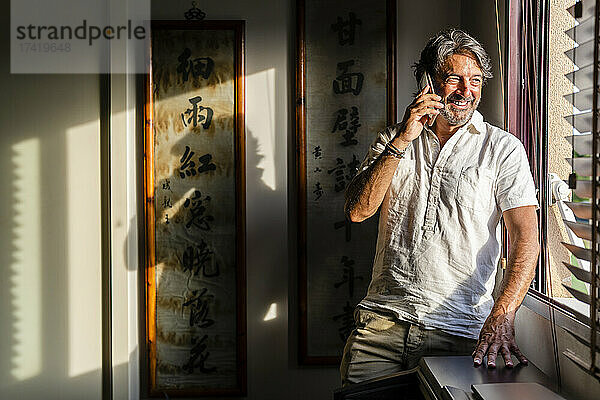 Lächelnder Mann  der durch das Fenster schaut  während er zu Hause mit dem Handy telefoniert