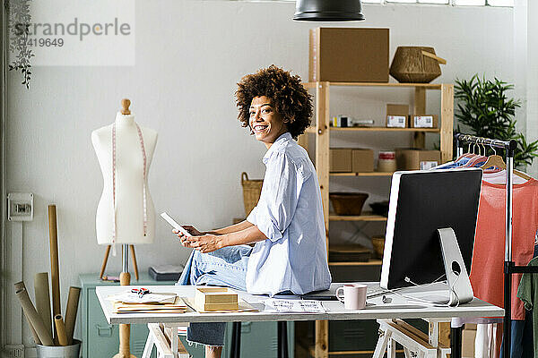 Lächelnde Designerin mit digitalem Tablet sitzt auf dem Schreibtisch im Studio