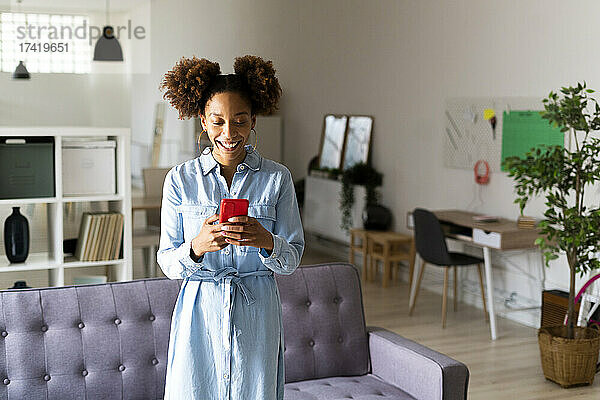 Glückliche junge Frau  die ihr Mobiltelefon benutzt  während sie im Wohnzimmer steht