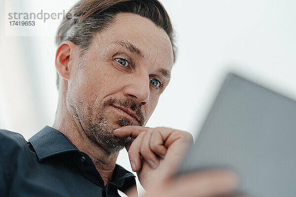 Geschäftsmann mit Hand am Kinn blickt im Büro auf digitales Tablet