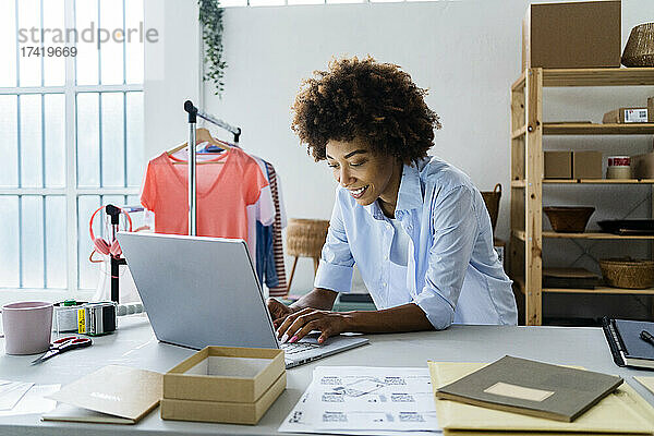 Junge Modedesignerin benutzt Laptop  während sie sich im Studio auf den Schreibtisch lehnt