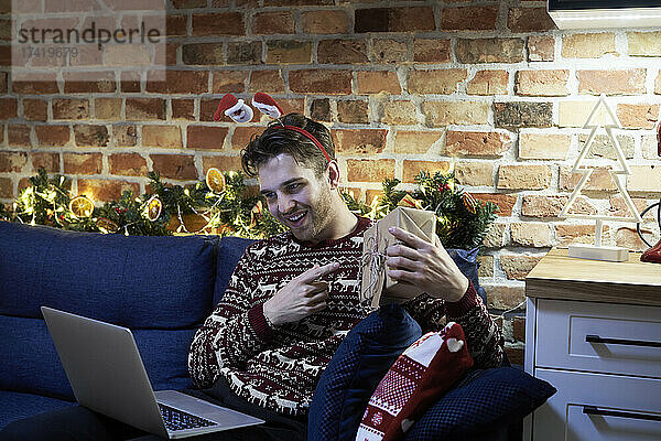 Lächelnder Mann zeigt Geschenk während Videoanruf über Laptop im Wohnzimmer