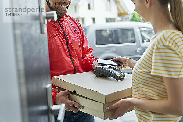 Frau bezahlt per Mobiltelefon  während sie vor der Tür eine Pizzabestellung vom Lieferboten entgegennimmt