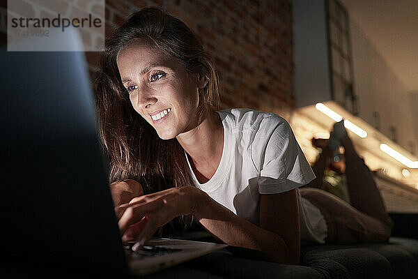 Junge Frau benutzt Laptop  während sie nachts zu Hause auf dem Sofa liegt