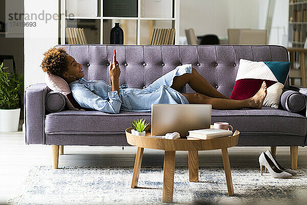 Lächelnde junge Frau benutzt Smartphone  während sie zu Hause auf dem Sofa liegt