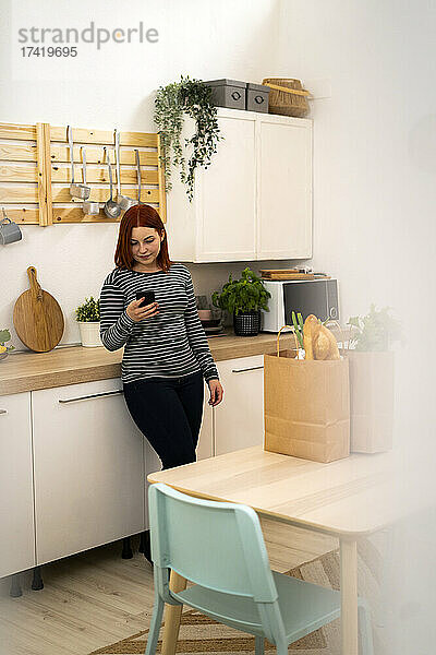 Rothaarige Frau benutzt Mobiltelefon  während sie in der Küche steht