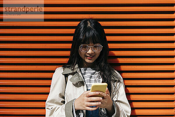 Junge Frau mit Brille und Smartphone vor orangefarbener Wand
