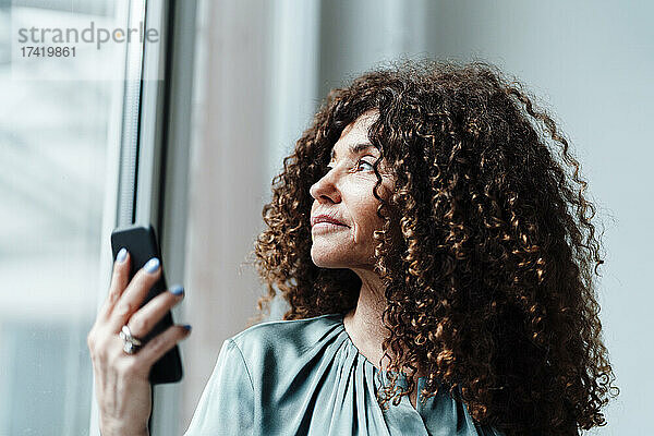 Geschäftsfrau blickt durch Glas  während sie ihr Smartphone in der Hand hält