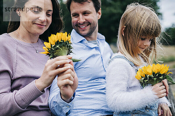 Frau und Mädchen halten Sonnenblume in der Hand  während sie mit Mann im Park sitzen