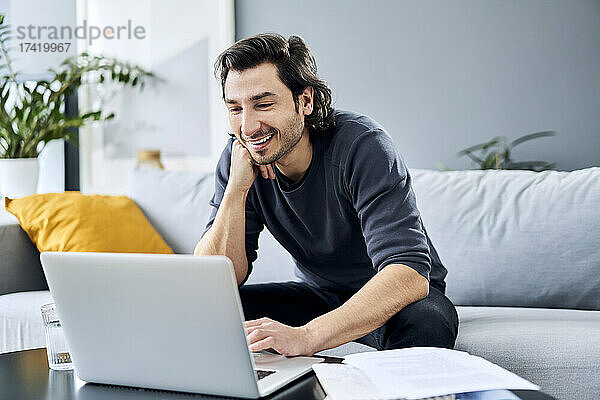 Lächelnder Geschäftsmann mit der Hand am Kinn  der zu Hause einen Laptop benutzt
