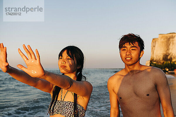 Junger Mann steht im Urlaub neben Frau und gestikuliert am Strand