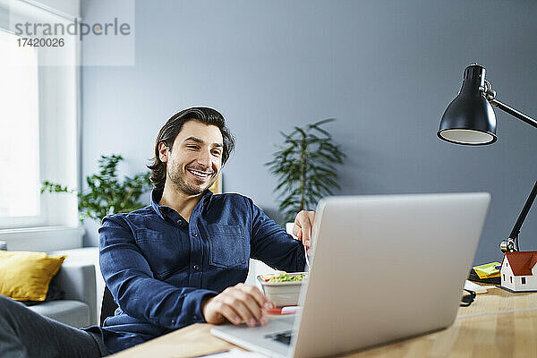 Lächelnder Geschäftsmann benutzt Laptop am Schreibtisch  während er im Büro sitzt