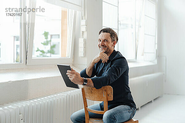 Lächelnder Geschäftsmann hält digitales Tablet in der Hand  während er auf einem Stuhl im Büro sitzt