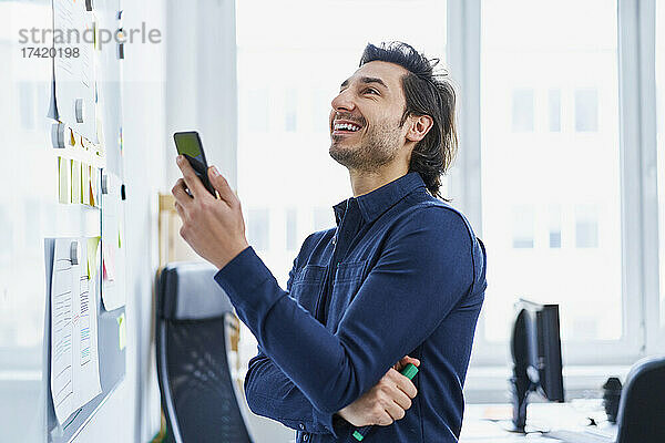 Lächelnder männlicher Webdesigner blickt auf das Whiteboard  während er im Büro sein Smartphone in der Hand hält