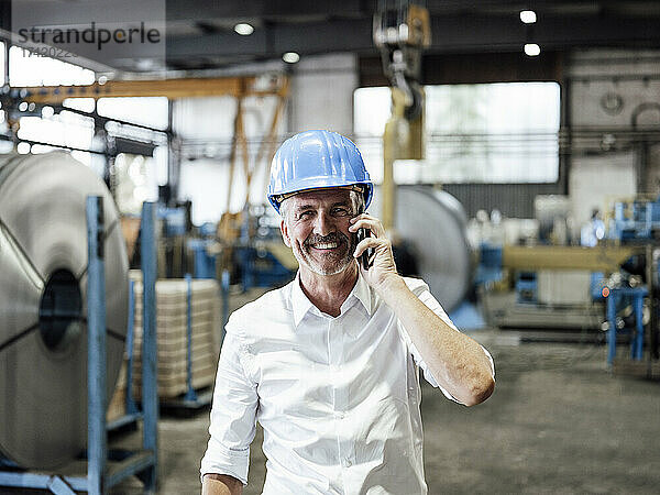 Lächelnder männlicher Ingenieur  der in der Metallindustrie mit dem Mobiltelefon spricht