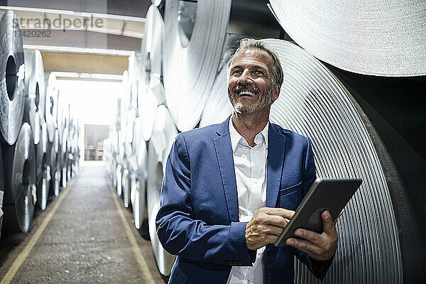 Lächelnder Geschäftsmann mit digitalem Tablet  der sich im Lager auf eine Blechrolle stützt