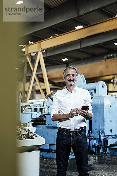 Lächelnder männlicher Geschäftsprofi mit Mobiltelefon  der im Stahlwerk steht