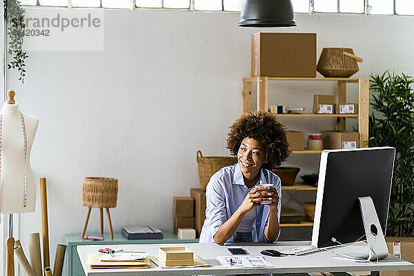 Lächelnde weibliche Designerin hält einen Becher in der Hand  während sie am Schreibtisch im Studio sitzt