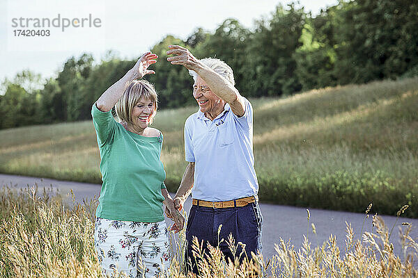 Glückliches älteres Paar mit erhobenen Händen tanzend  während es auf der Wiese steht
