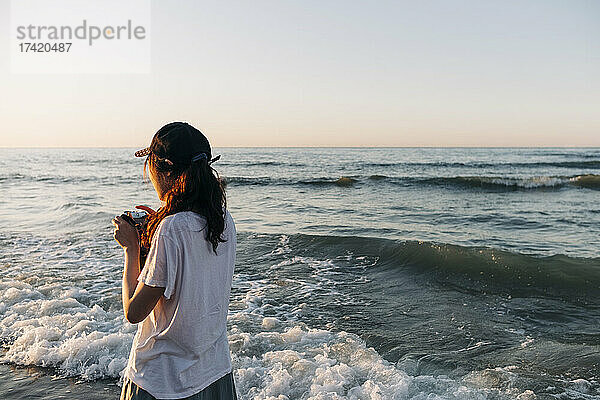 Junge Frau mit Kamera blickt im Urlaub aufs Meer