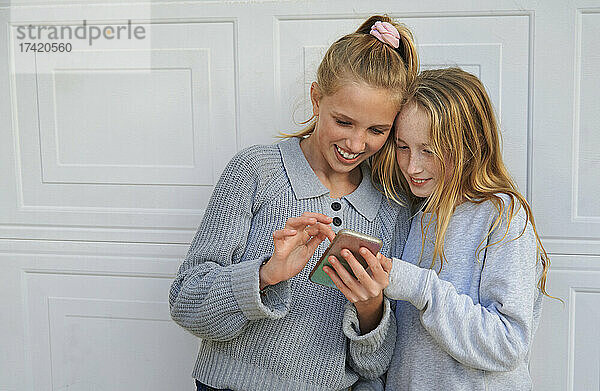Lächelnde Mädchen teilen ihr Mobiltelefon vor dem Garagentor