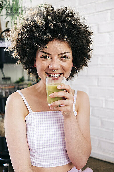 Lächelnde junge Frau hält ein Smoothie-Glas in der Hand  während sie zu Hause sitzt