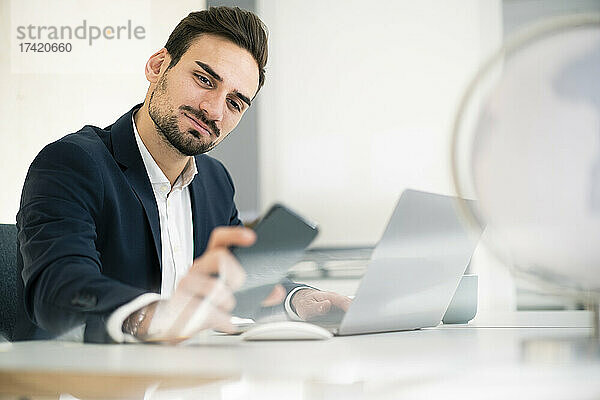 Schöner männlicher Profi  der sein Smartphone überprüft  während er mit Laptop am Schreibtisch im Büro sitzt