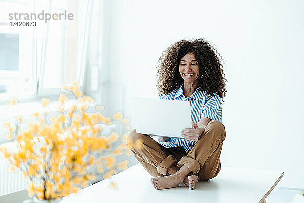 Lächelnde reife Geschäftsfrau benutzt Laptop  während sie mit gekreuzten Beinen am Tisch im Büro sitzt