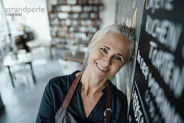 Lächelnde Cafébesitzerin mit Schürze lehnt an der Tafel im Café