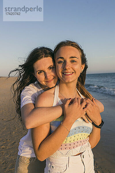 Lächelnde junge Frau umarmt ihre Freundin am Strand bei Sonnenuntergang