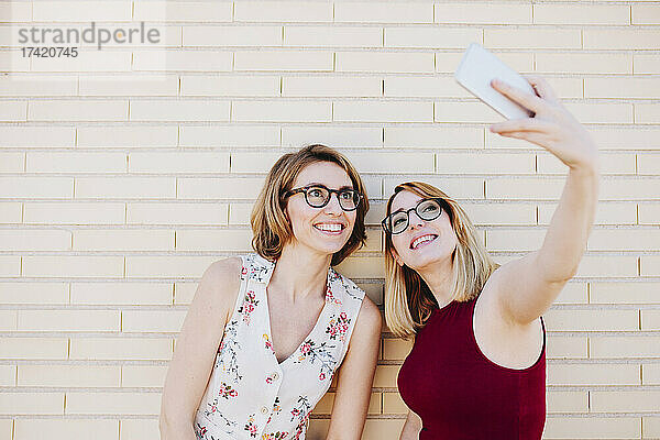 Lächelnde Kolleginnen machen ein Selfie mit dem Smartphone vor einer Ziegelwand
