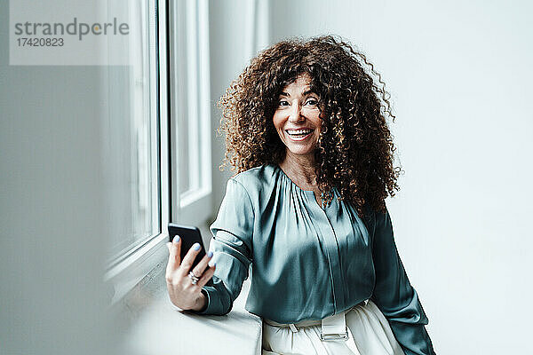 Lächelnde reife Geschäftsfrau mit Smartphone lehnt auf der Fensterbank