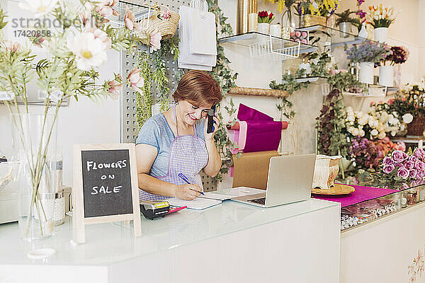 Lächelnde Blumenladenbesitzerin  die mit dem Smartphone spricht  während sie an der Kasse Tagebuch schreibt