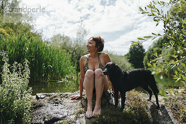 Lächelnde Frau sitzt mit Hund auf einem Felsen an einem Teich